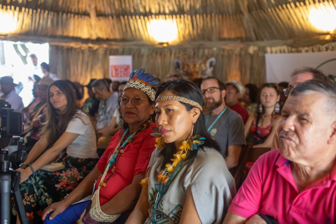 Mujeres indígenas con diademas y plumas escuchando atentas dentro de una maloca