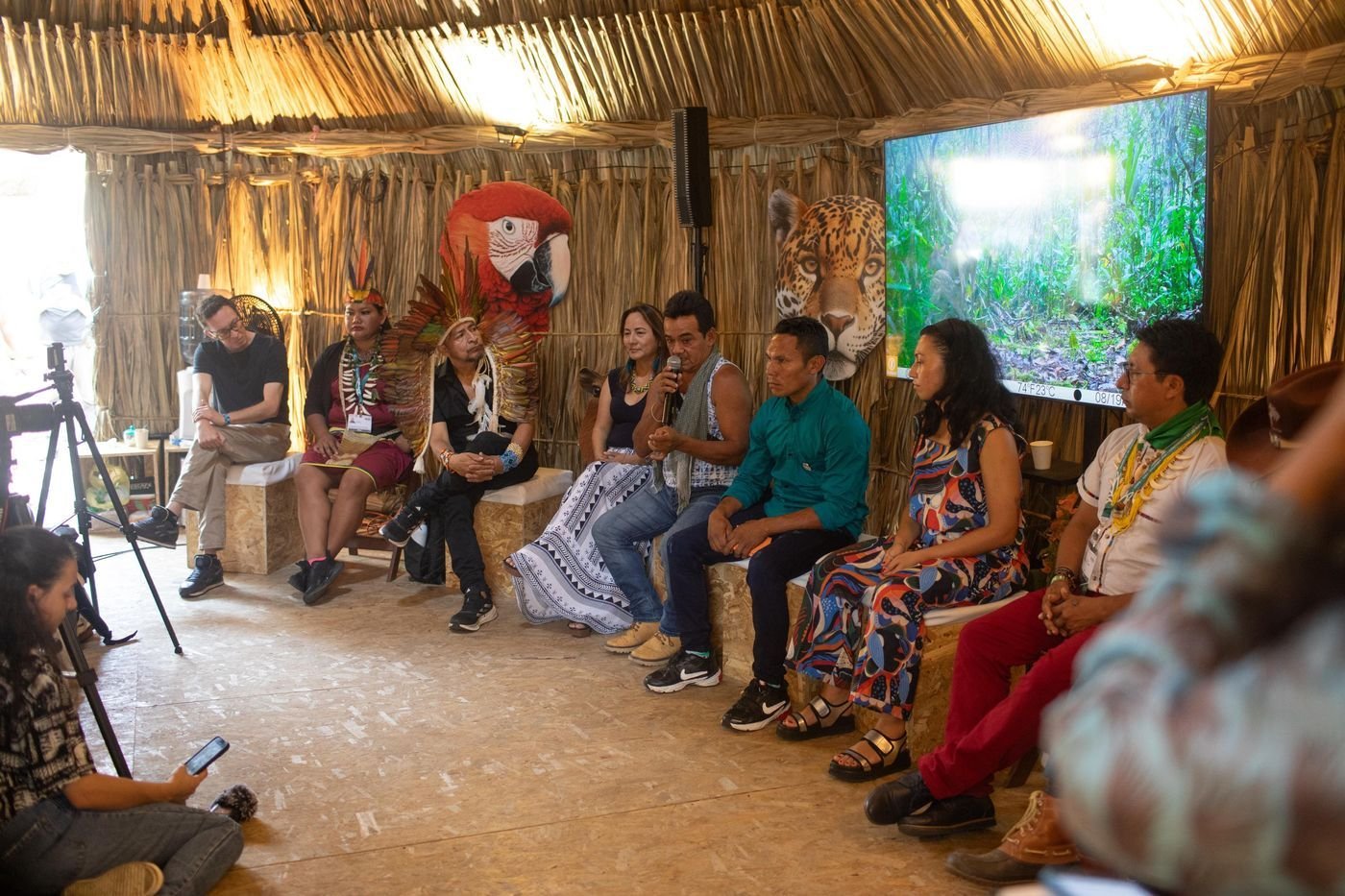 Panel de personas sentadas en círculo dentro de una maloca con un guacamayo y un jaguar pintados al fondo