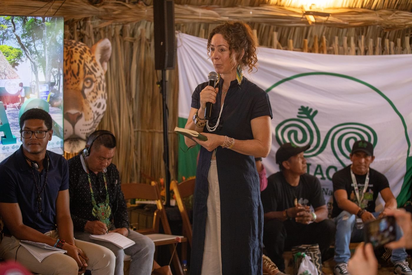 Mujer de pie hablando con micrófono frente a una bandera blanca con espirales verdes