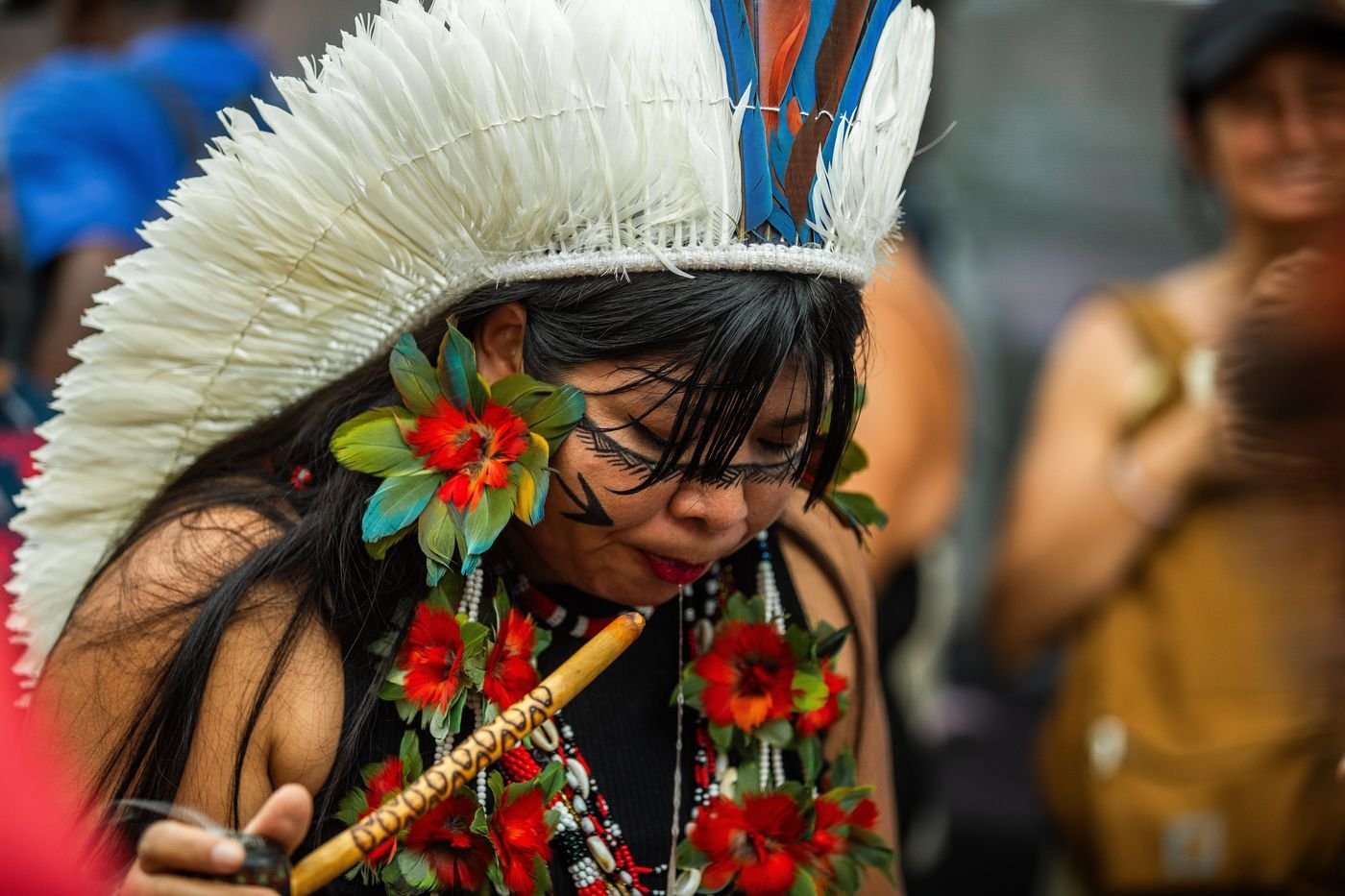 Mujer indígena con gran tocado de plumas blancas y rostro pintado tocando un instrumento