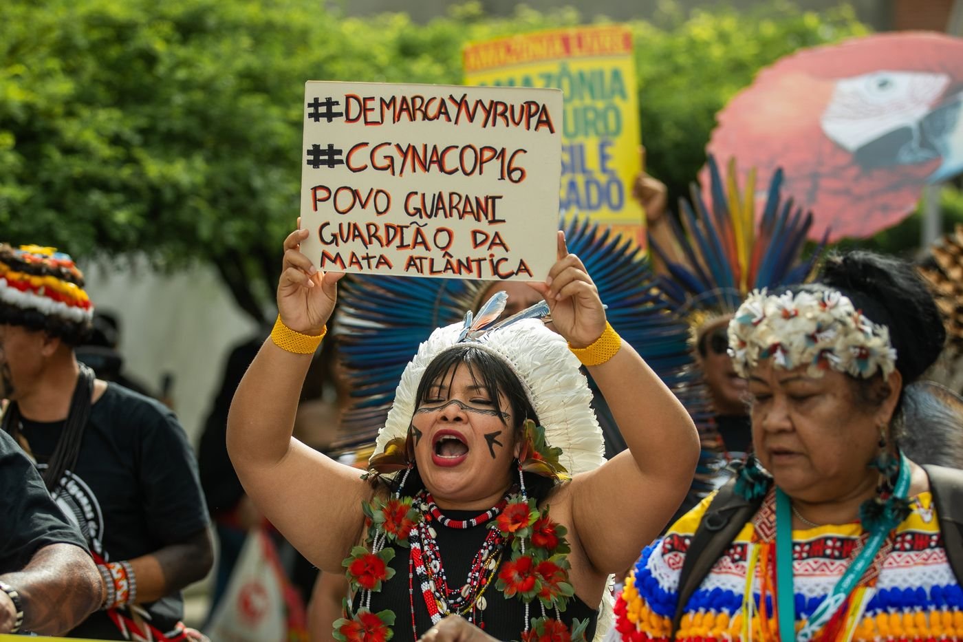 Mujer indígena con tocado de plumas blancas sostiene un cartel durante una marcha