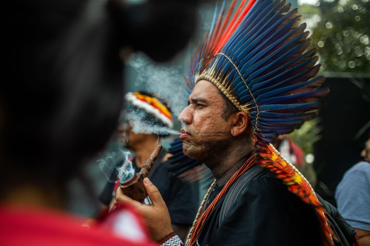 Hombre indígena con un gran tocado de plumas azules y rojas soplando humo de una pipa